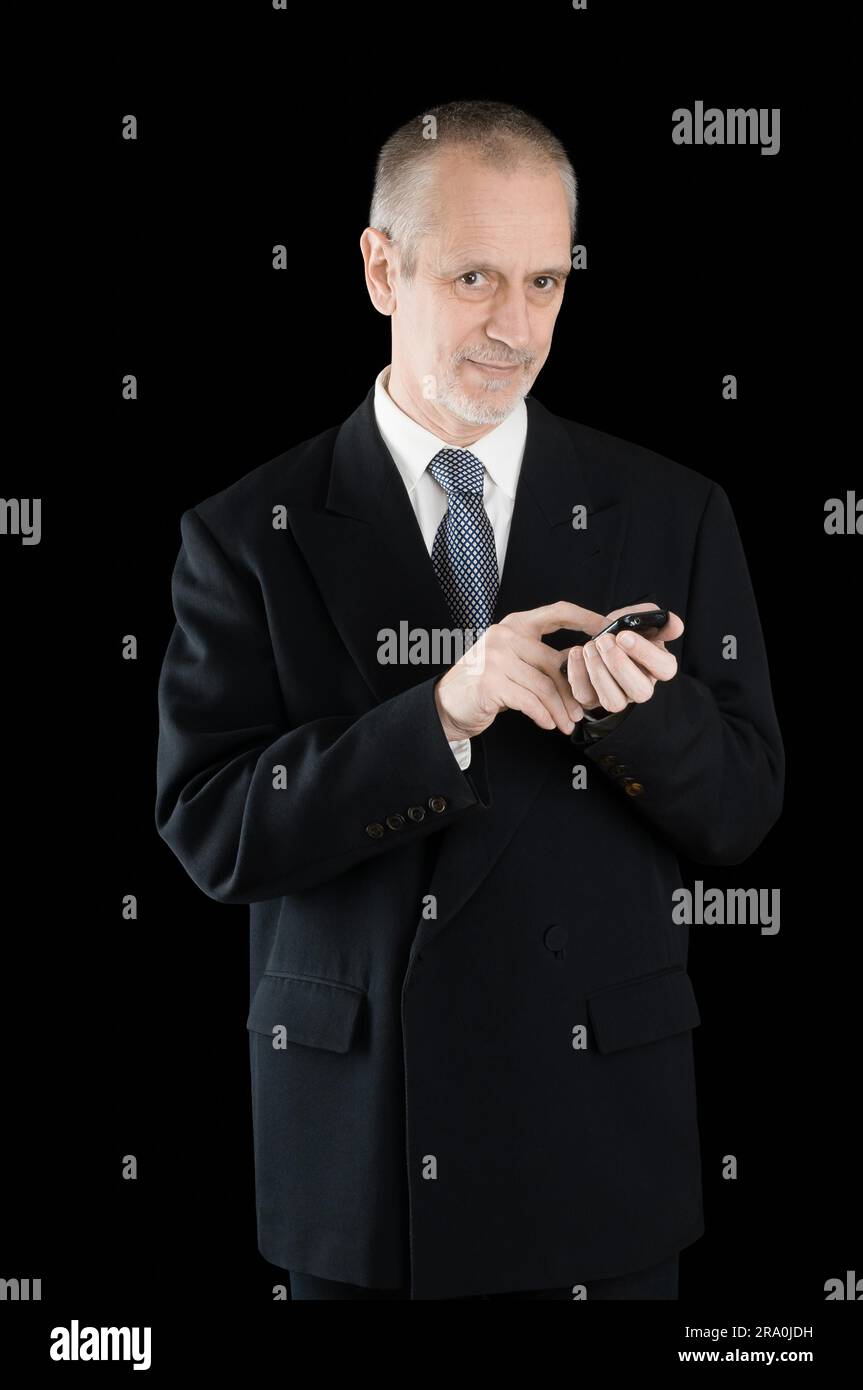 Imprenditore Senior in abito nero, la scrittura di sms o comporre un numero sul suo telefono cellulare, su sfondo nero Foto Stock