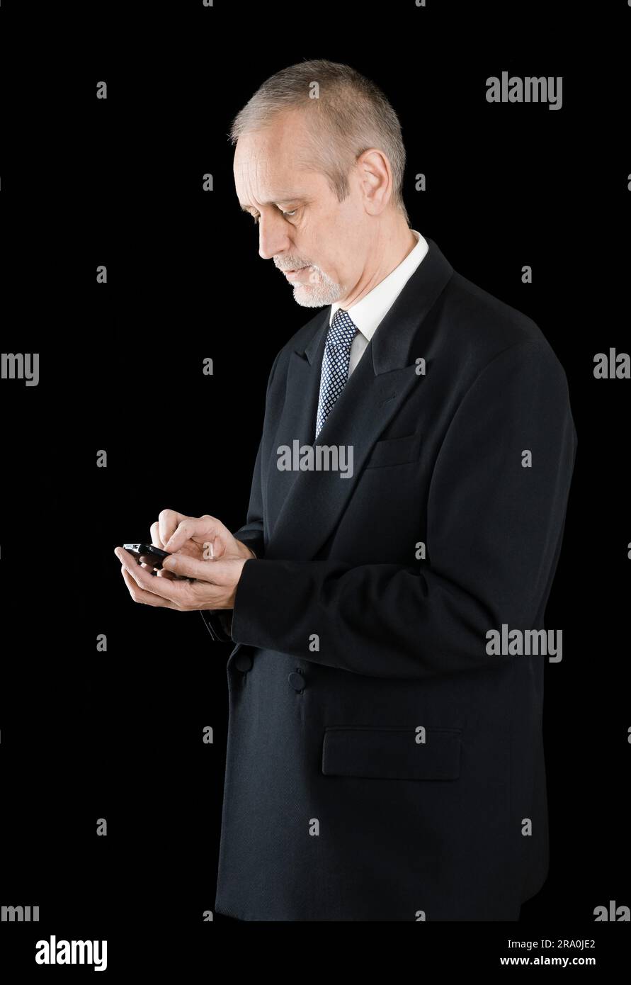 Imprenditore Senior in abito nero la scrittura di sms o comporre un numero sul suo telefono cellulare, su sfondo nero Foto Stock