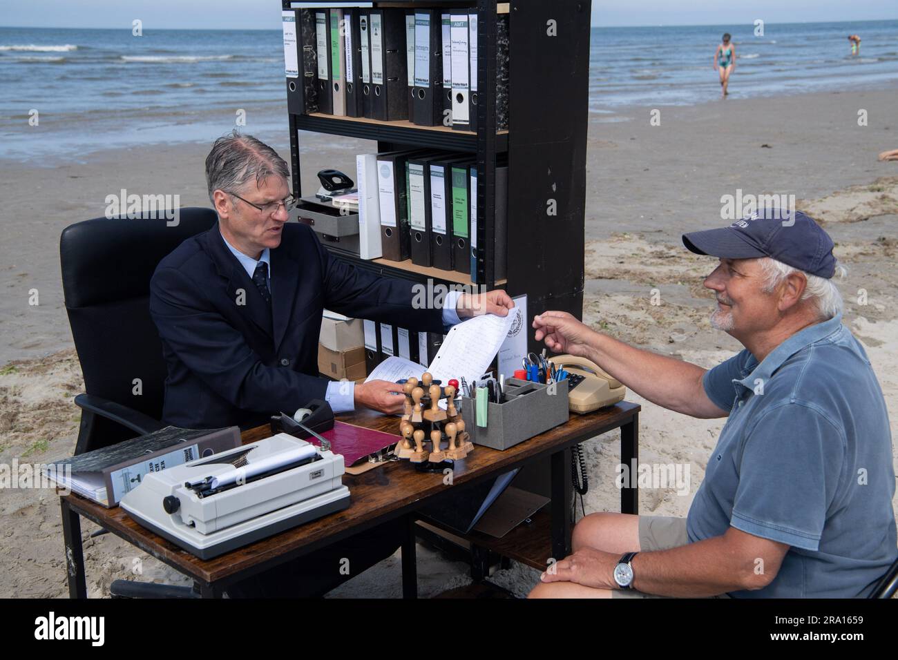 Spiekeroog, Germania. 29 giugno 2023. Albrecht Fersch (l) siede alla sua scrivania sulla spiaggia dell'isola orientale frisone di Spiekeroog e parla con il turista Falko Grunau. Con il suo progetto "Schreibtischtäter", l'artista d'azione vuole usare l'umorismo per incoraggiare le persone a pensare alle condizioni sociali e all'esistenza umana. Crediti: Marco Rauch/dpa/Alamy Live News Foto Stock
