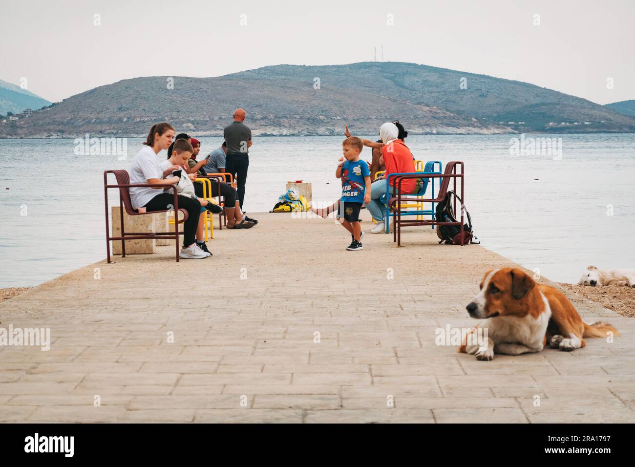 Un cane riposa mentre la gente siede sui sedili lungo un molo di cemento sulla spiaggia principale della città di Sarandë, nella riviera albanese Foto Stock