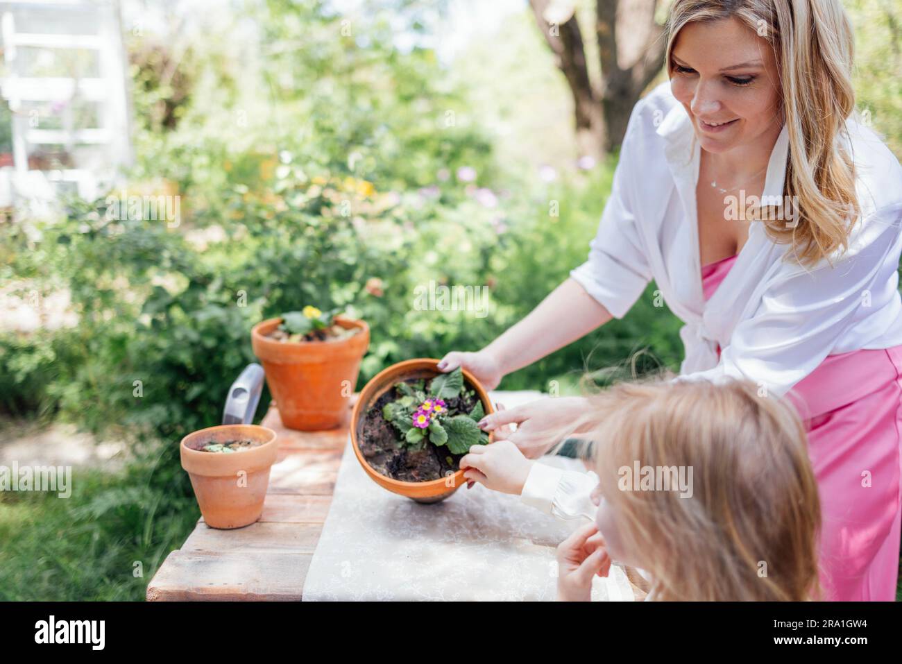 La bambina e la sua bella mamma bionda stanno piantando fiori in vasi di argilla all'aperto in giardino. La giovane donna e sua figlia carina passano del tempo insieme in b Foto Stock