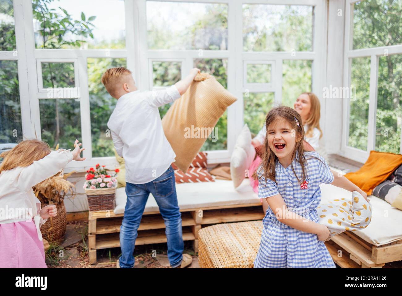 Una grande famiglia allegra si diverte nel gazebo di vetro in giardino. Tre bambini e una piccola e affascinante madre mettono cuscini e ridono nel cortile di casa. SM Foto Stock