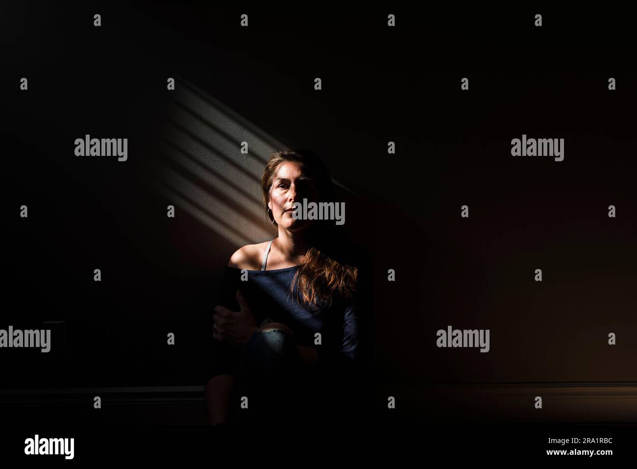 Ritratto di una donna seduta in una zona di luce a strisce in una stanza buia. Foto Stock