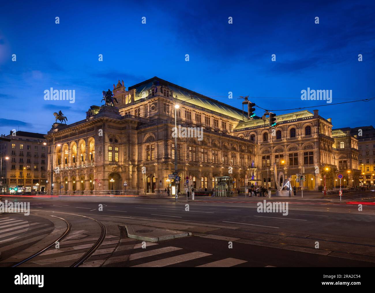 Teatro dell'Opera di Vienna illuminato durante l'ora blu/crepuscolo. Persone non identificabili a causa della sfocatura del movimento. Strisce leggere causate dal passaggio di tram e auto Foto Stock