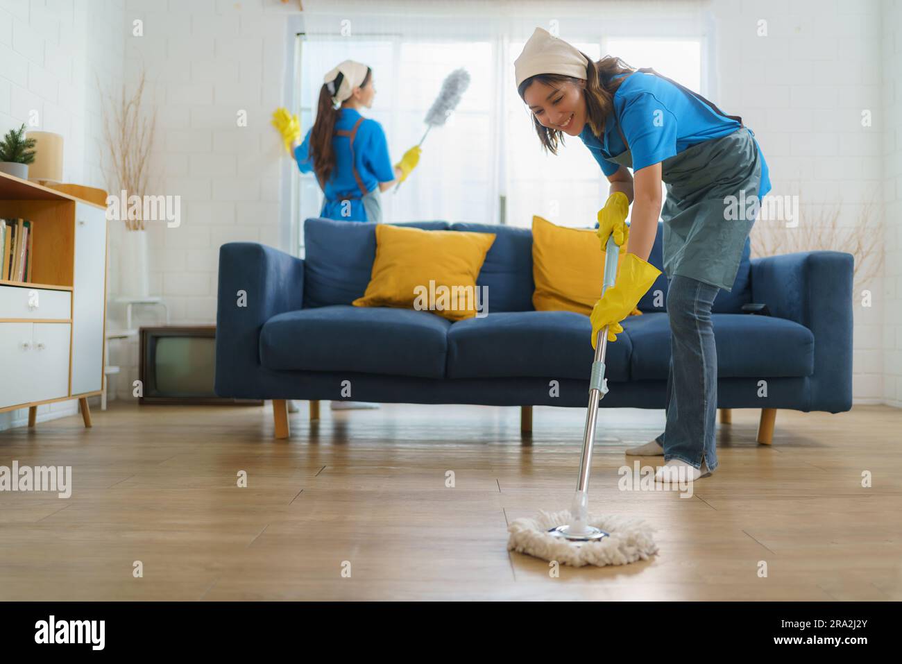 Due diligenti cameriere asiatiche collaborano senza problemi, pulendo il soggiorno con precisione ed efficienza, senza lasciare alcun angolo intatto nella loro ricerca Foto Stock