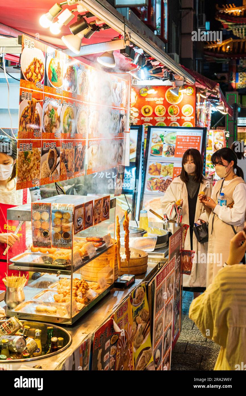 Popolare Chinatown a Kobe, Giappone. Chiosco di fast food cinese che vende gnocchi e panini cotti al vapore, 2 giovani donne che mangiano mentre guardano il bancone del cibo. Foto Stock