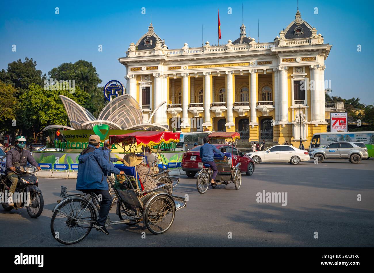 Cicloi (pedicabs) che trasportano turisti e altri passi di traffico di fronte al teatro dell'opera di Hanoi dell'epoca coloniale francese, o Nha Hat Lon, ad Hanoi, in Vietnam. Foto Stock