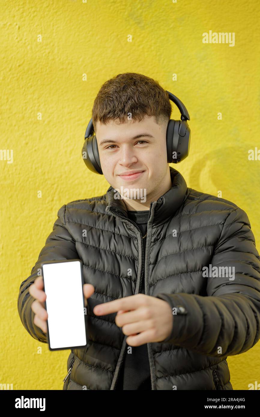 Il ragazzo caucasico con le cuffie mostra lo schermo vuoto del suo telefono cellulare isolato su uno sfondo giallo. Foto Stock