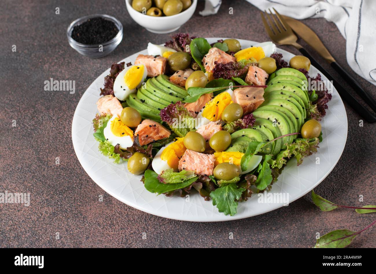 Insalata con salmone al forno, avocado, uova e olive, cosparsa di semi di sesamo su un piatto bianco su sfondo marrone Foto Stock