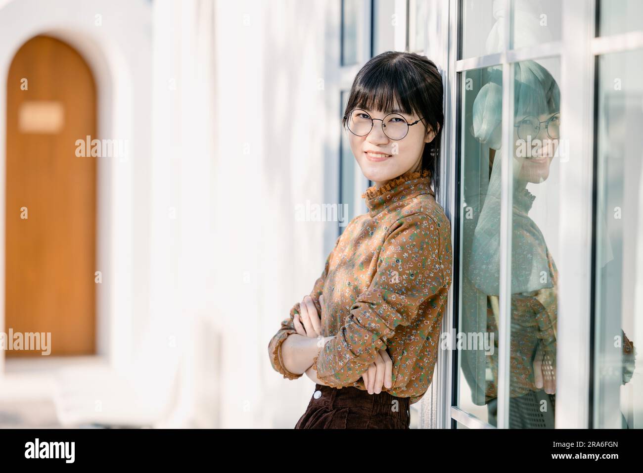 Ritratto giovane occhiali sottili donna sorridente che guarda la fotocamera a braccia incrociate. Ragazza felice in piedi sullo sfondo dell'ufficio. Standi donna d'affari asiatica di successo Foto Stock