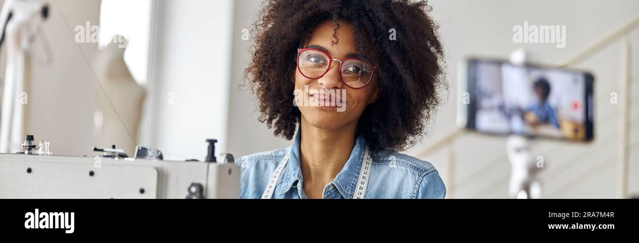 Bella donna afro-americana blogger con forbici e occhiali sul posto di lavoro in laboratorio di cucito Foto Stock