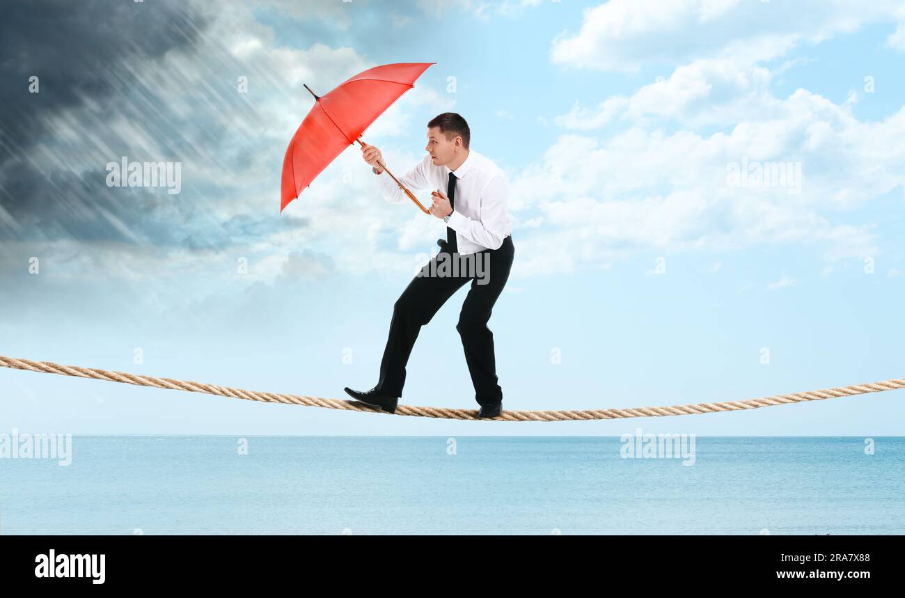 Rischi e sfide dell'imprenditorialità. Uomo d'affari con ombrello che protegge dalle precipitazioni in corda sul mare Foto Stock