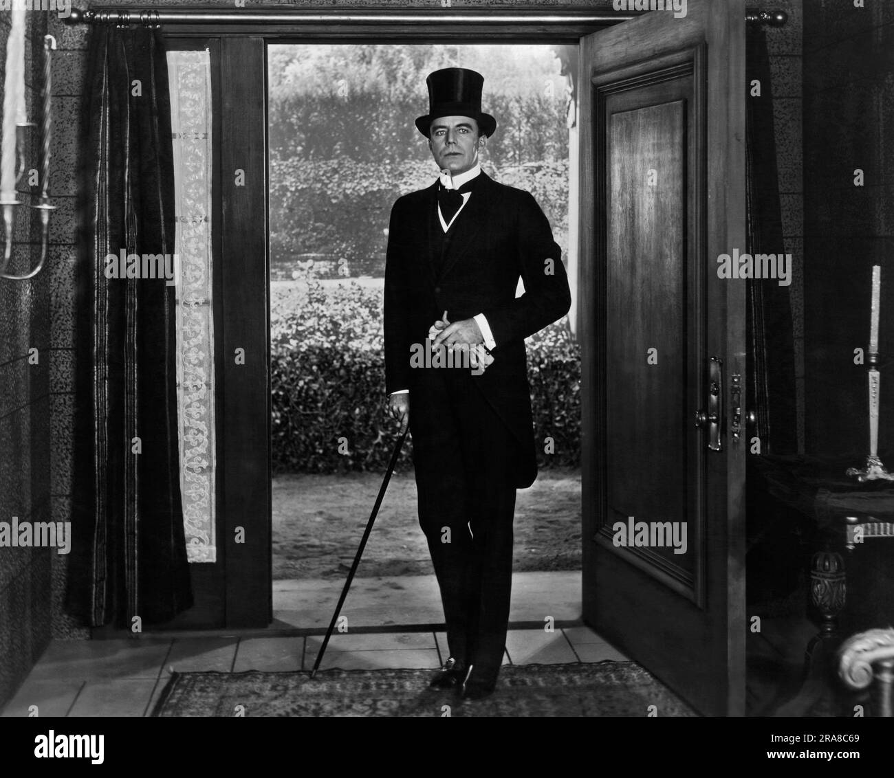 Hollywood, California, c 1918 la star del cinema silenzioso e attore House Peters entra in una casa indossando uno smoking, un cappello e portando un bastone. Foto Stock