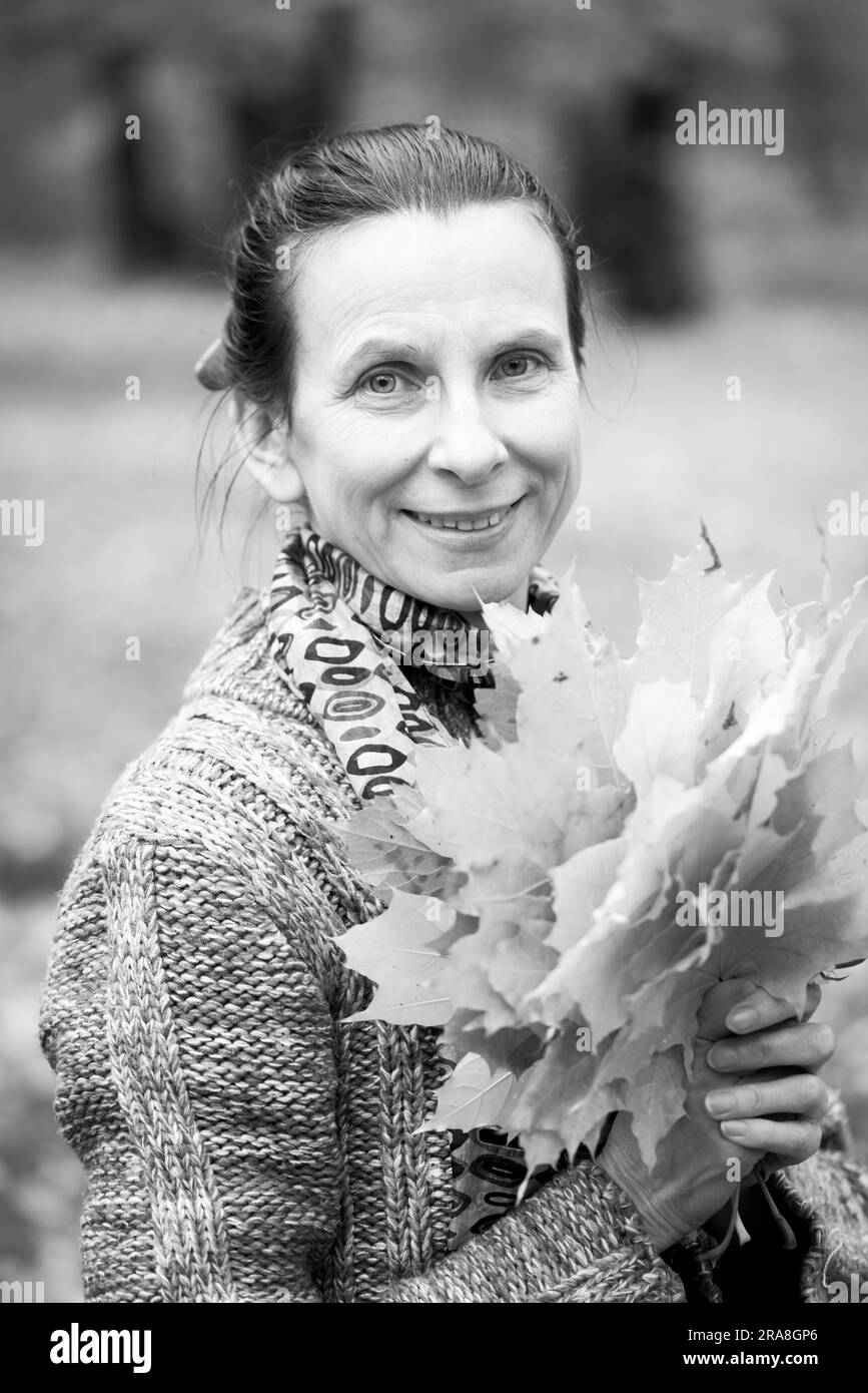 Ritratto in bianco e nero di un sorridente adulto donna caucasica è raccolta giallo foglie di acero in autunno nel parco Foto Stock