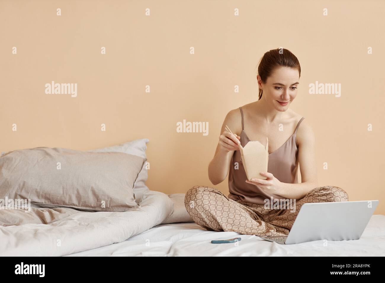 Ritratto minimale di una giovane donna sorridente che mangia spaghetti da asporto e guarda video tramite computer portatile, mentre ti godi un weekend rilassante a casa e copia Foto Stock