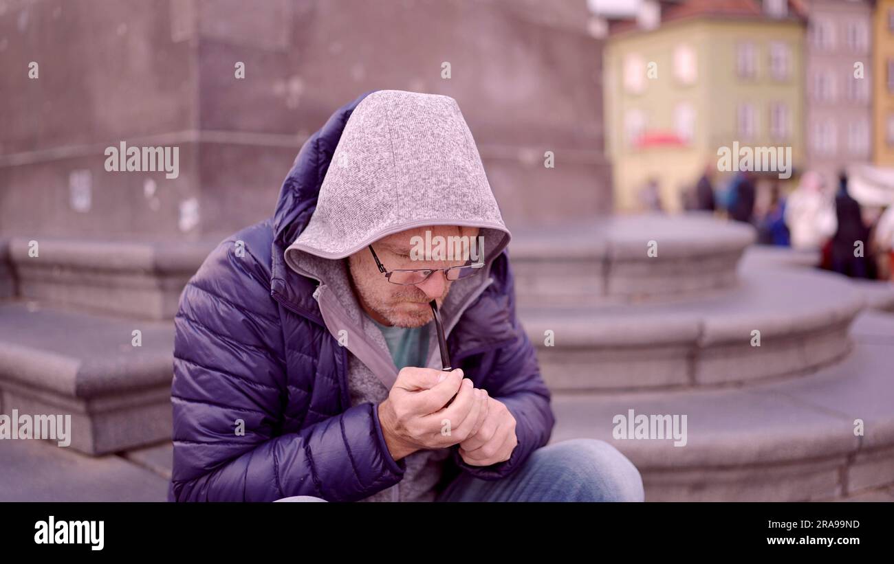Un uomo adulto в очках seduto sulla piazza si illumina davanti a una pipa da tabacco in Piazza del Palazzo, nella città vecchia di Varsavia Foto Stock