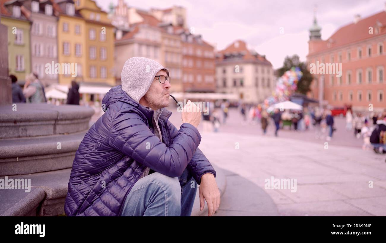 Ritratto di un uomo adulto con occhiali seduti nel cappuccio sulla piazza e fumare un tubo di tabacco nella Piazza del Palazzo, nella Città Vecchia di Varsavia Foto Stock
