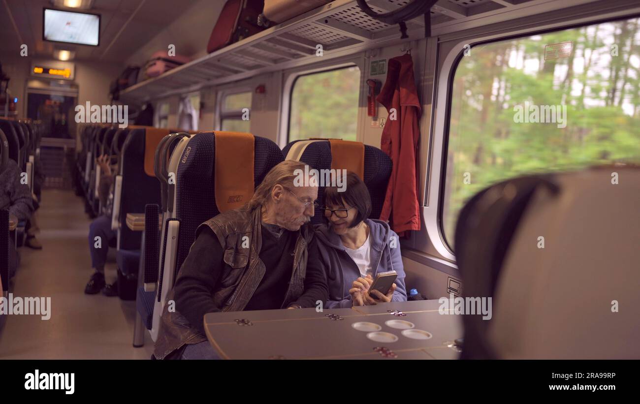 La coppia anziana viaggia in treno, la signora tiene in mano un telefono cellulare, entrambi guardano lo smartphone e parlano tra loro Foto Stock
