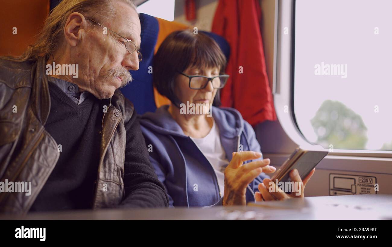 La coppia anziana viaggia in treno, la signora tiene in mano un telefono cellulare, entrambi guardano lo smartphone e parlano tra loro Foto Stock