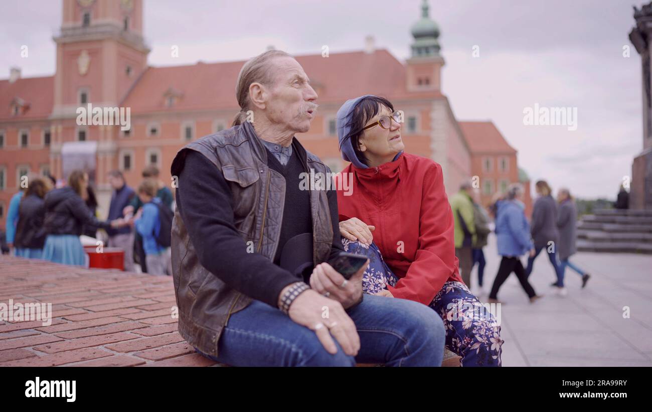Una coppia di turisti anziani è seduta e parla nel centro storico di una vecchia città europea, senior ha uno smartphone in mano. Piazza del Palazzo, Foto Stock