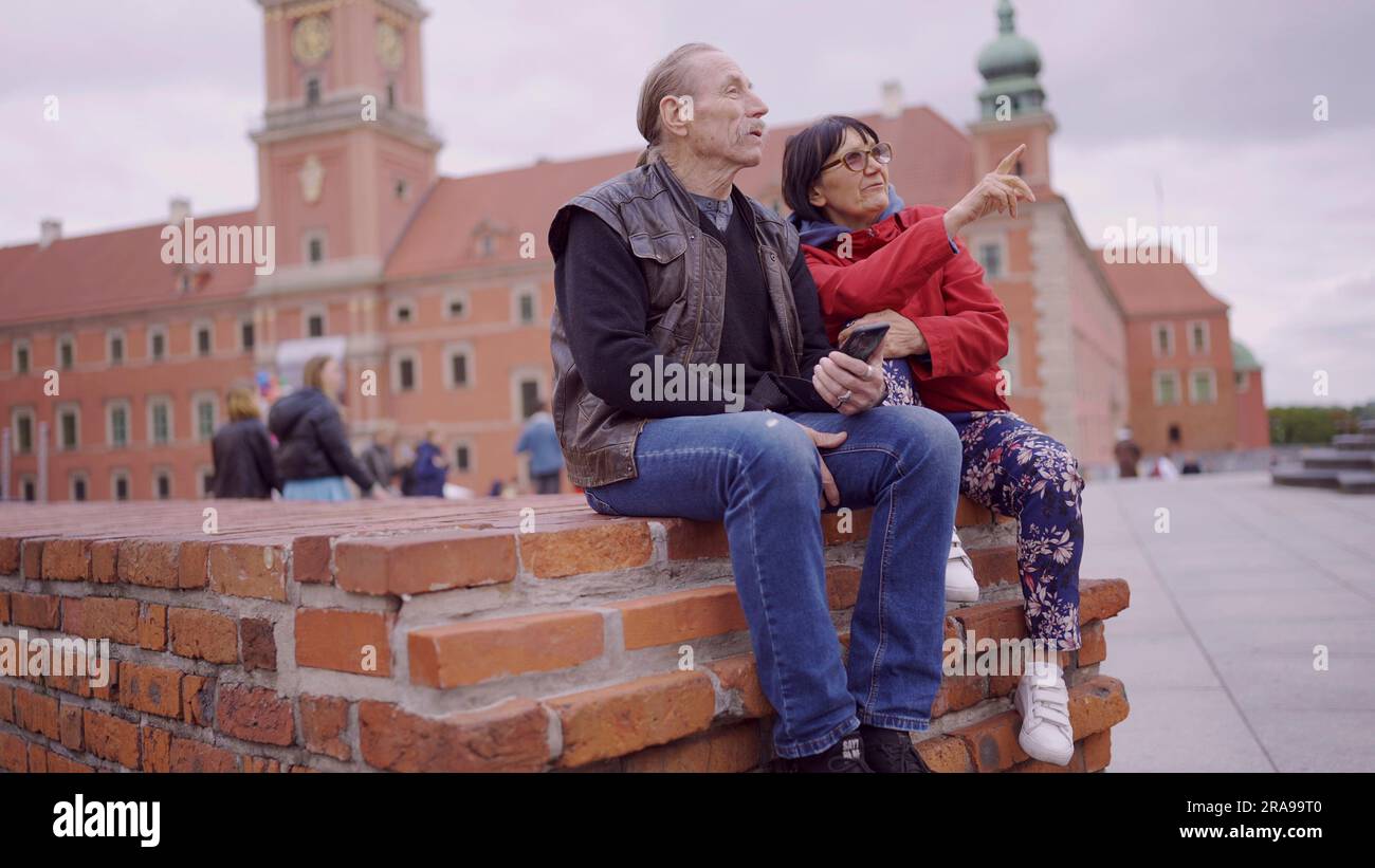 Una coppia di turisti anziani è seduta e parla nel centro storico di una vecchia città europea, senior ha uno smartphone in mano. Piazza del Palazzo, Foto Stock