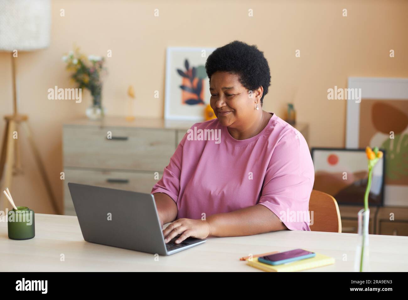 Donna afro-americana matura e felice con una t-shirt rosa seduta accanto alla scrivania di fronte al laptop in salotto, digitando sulla tastiera e collegando in rete Foto Stock