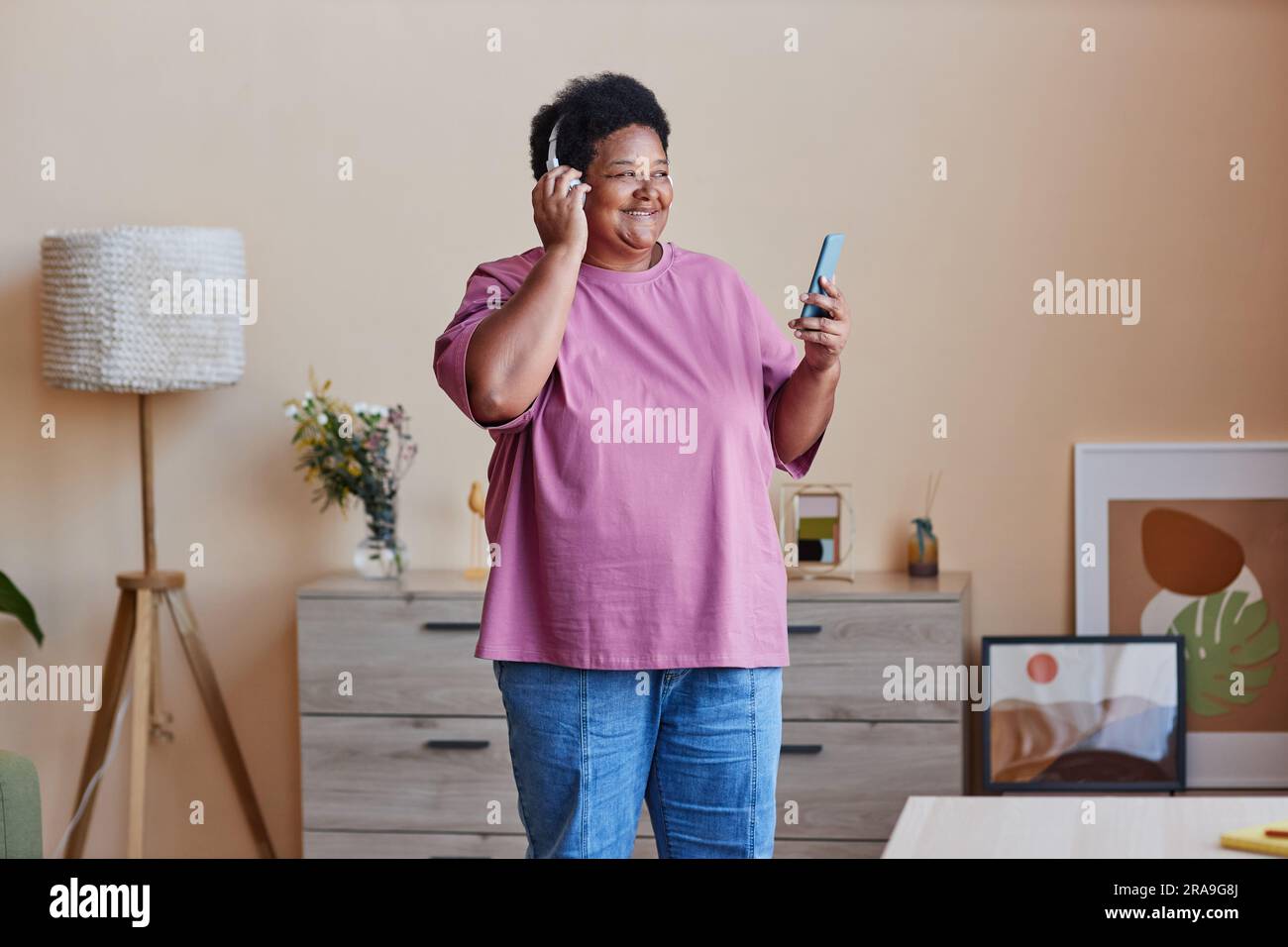 Donna afro-americana anziana e allegra con smartphone che ascolta la sua musica preferita in cuffia o guarda video o film online Foto Stock