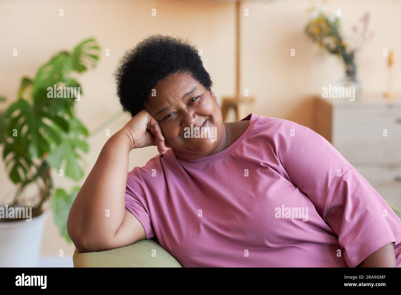 Donna anziana afro-americana felice con t-shirt rosa seduta in poltrona davanti alla macchina fotografica e guardata con sorriso in ambiente domestico Foto Stock
