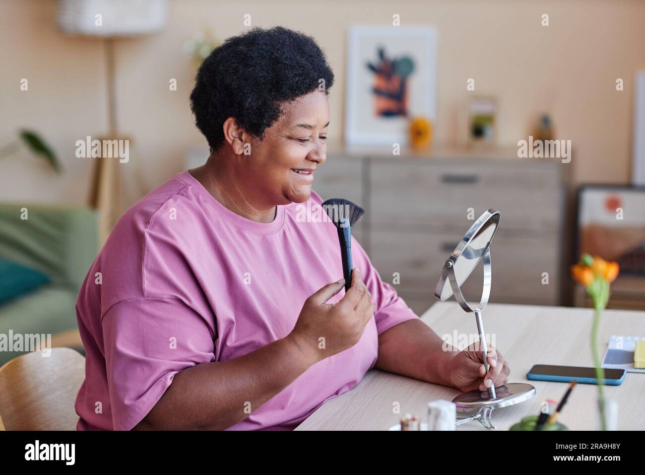 Donna afro-americana matura felice che guarda allo specchio mentre si siede accanto al tavolo in salotto e si applica della polvere sul viso Foto Stock