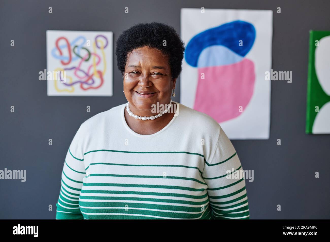 Felice insegnante d'arte maturo in abbigliamento casual davanti alla macchina fotografica contro opere d'arte appese a una parete grigia dello studio e che ti guarda Foto Stock