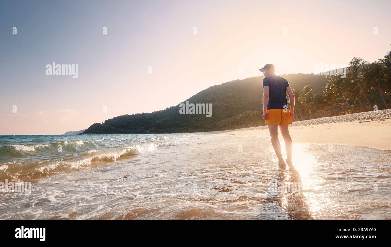 Vista posteriore di Happy Man mentre cammini in mare. Viaggiatore solo su un'idilliaca spiaggia di sabbia vuota con palme al tramonto. Isole Cham, Vietnam. Foto Stock