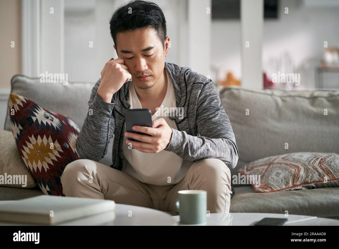 giovane asiatico seduto sul divano a casa guardando il cellulare sembra serio Foto Stock