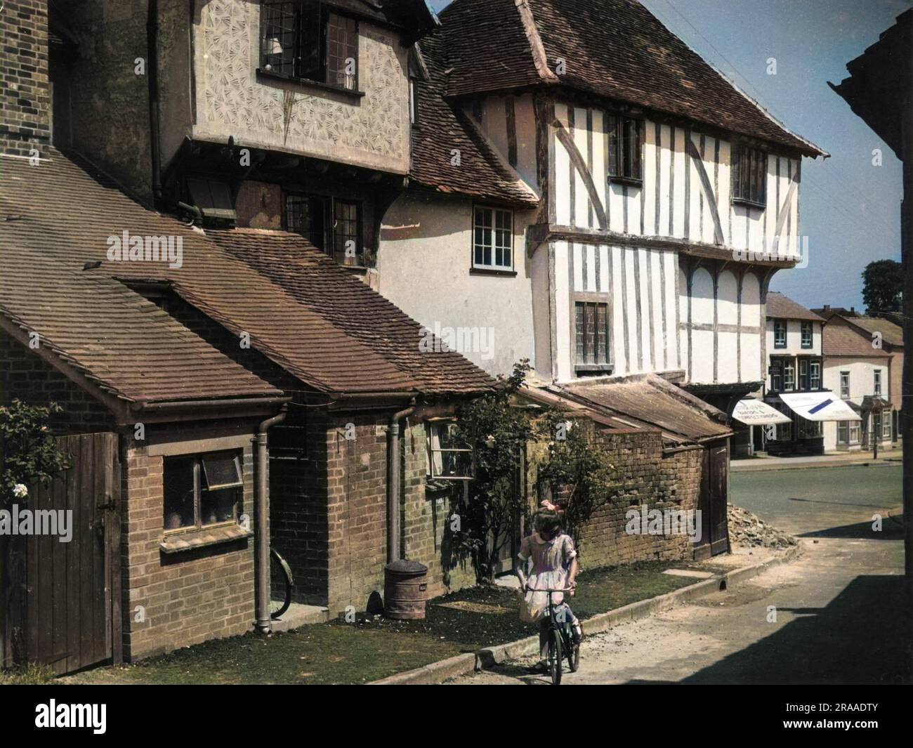 Una bambina con un fiocco tra i capelli cavalca la sua bicicletta in un angolo della pittoresca città vecchia di Thaxted, Essex, Inghilterra, con i suoi vecchi cottage con tetto in paglia accanto alla Guild Hall Data: Anni '1950 Foto Stock