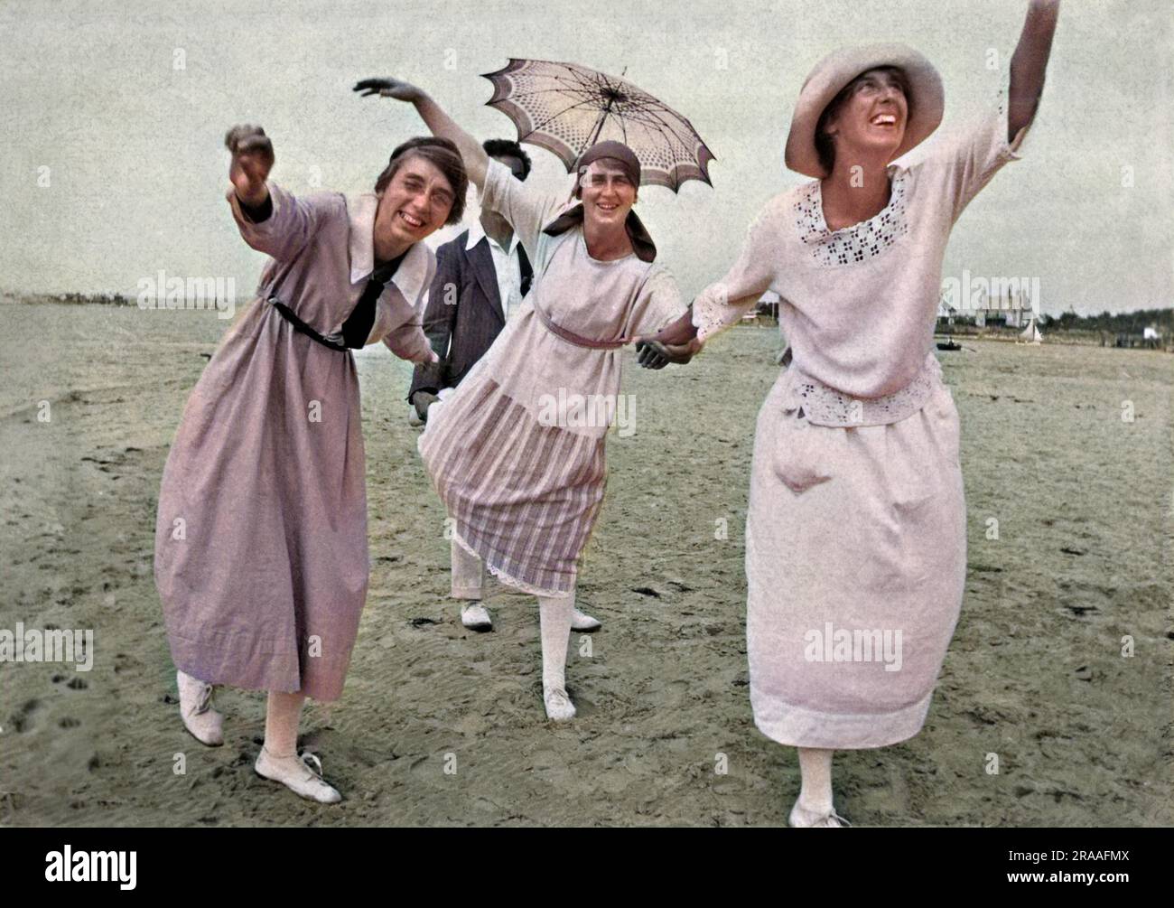 Tre donne che colpiscono pose divertenti su una spiaggia. Un uomo sullo sfondo tiene un ombrellone. Foto Stock