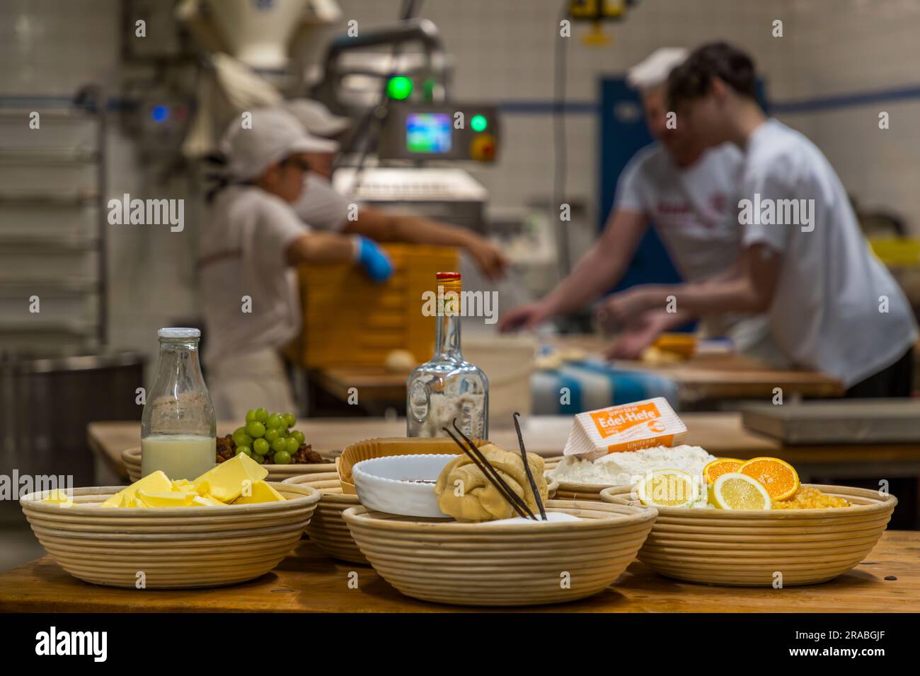 Ingredienti per il Christstollen originale di Dresda disposti in cesti da panettiere su un tavolo da lavoro: burro, uva verde, una bottiglia di rum, farina e fette di limone e arancia sullo zucchero candito. sullo sfondo, il personale della panetteria lavora a un impastatore. La fotografia è stata scattata presso la panetteria Dresdner Backhaus, Dresda, Sassonia, Germania. Foto Stock