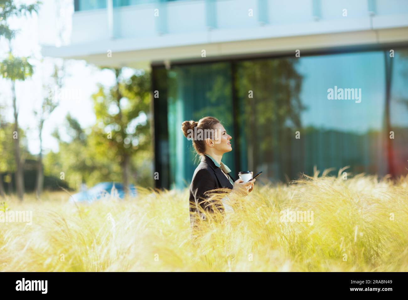 penosa donna d'affari moderna vicino all'edificio degli uffici, con giacca nera con smartphone e tazza di caffè. Foto Stock