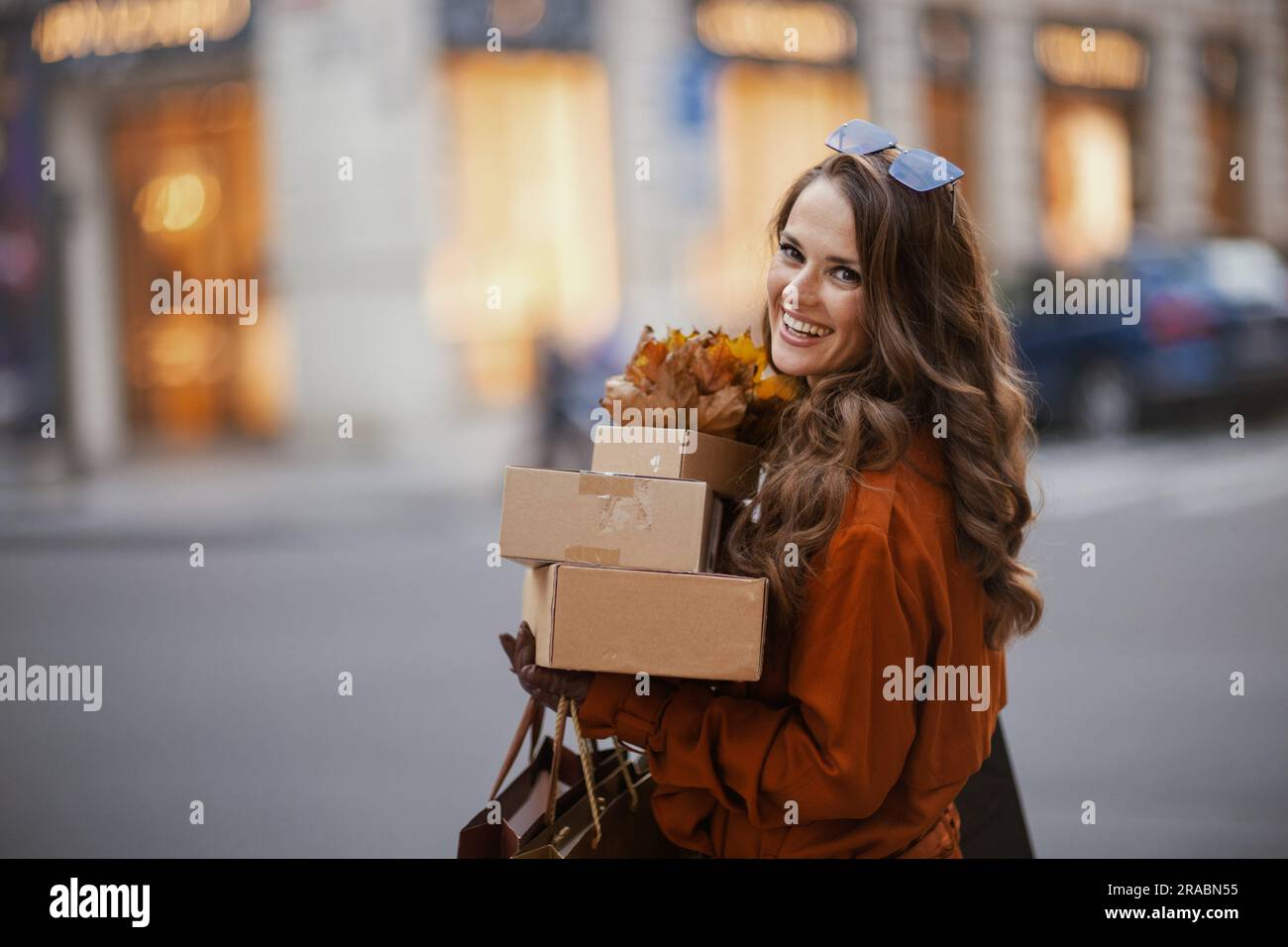 Ciao november. giovane donna sorridente in trench marrone con pacchi, borse della spesa e foglie gialle autunnali in città. Foto Stock