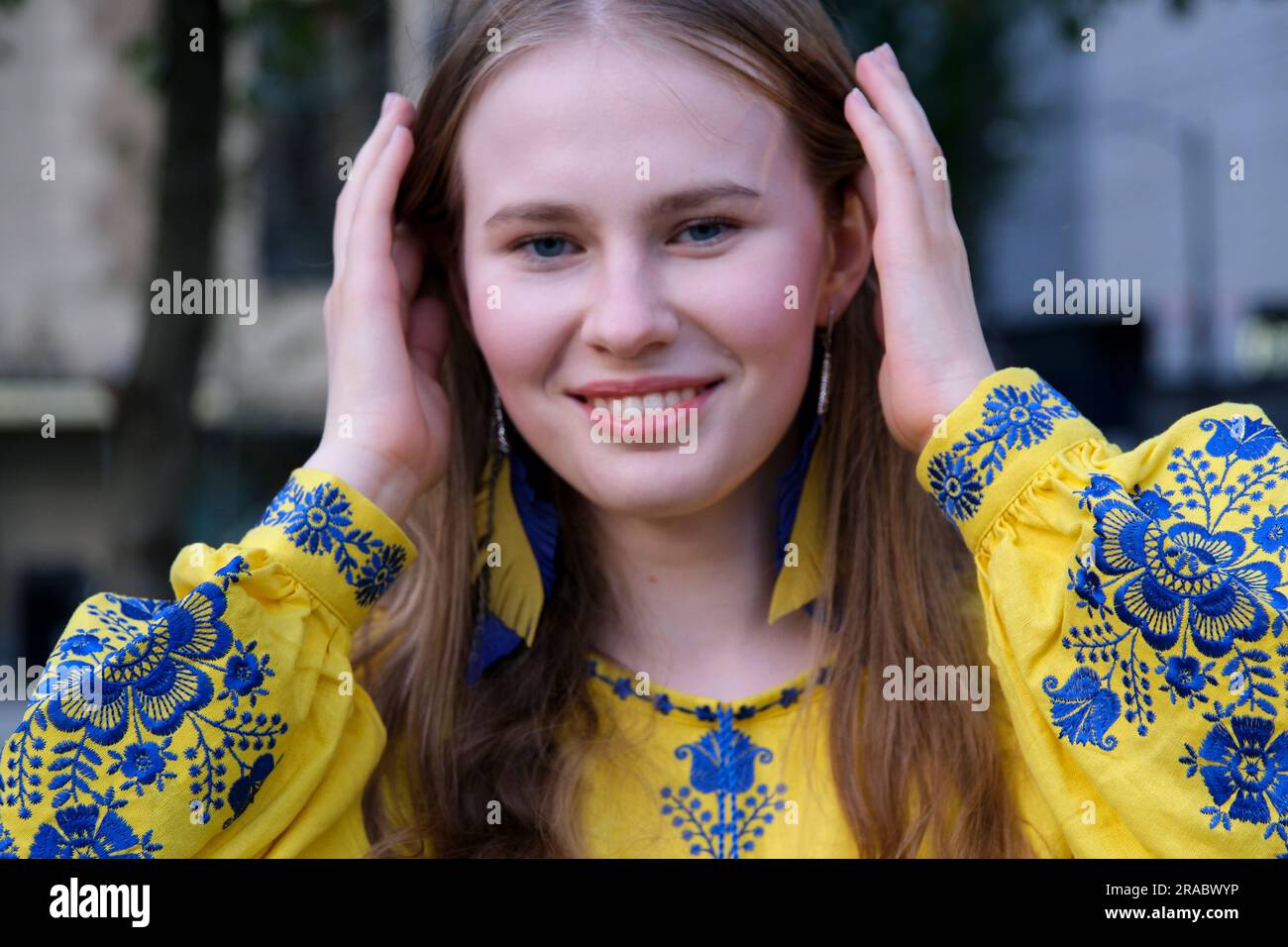 La camicia ricamata della giovane donna Ucraina guarda nelle raddrizzature dei sorrisi. Capelli biondi sfondo sfocato primi piani orecchini facciali sotto forma di bandiera dell'Ucraina divertimento della vittoria gioia forza libertà volontà Foto Stock