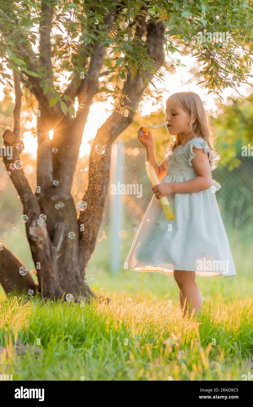 una bambina in un vestito di cotone estivo soffia bolle di sapone in estate al tramonto in un parco vicino a un albero, spazio per il testo. Foto di alta qualità Foto Stock