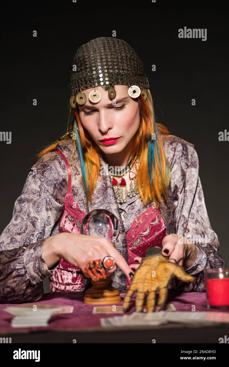 Seria redhead femmina indovina in costume e copricapo zingari che mostra la previsione del futuro su palme artificiali su sfondo scuro Foto Stock