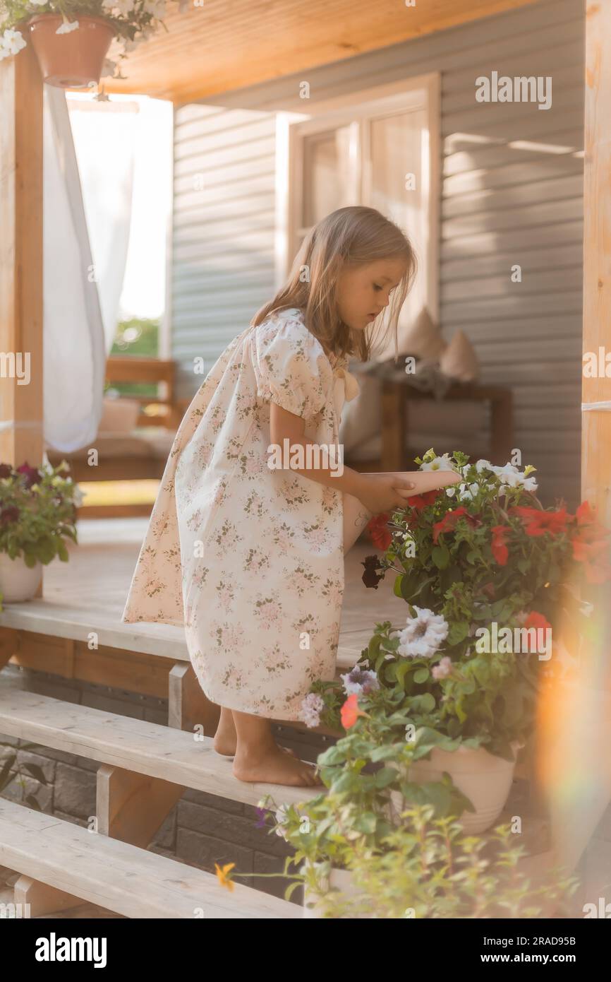 la bambina con i capelli biondi annaffia i fiori sulla veranda d'estate al tramonto. Foto di alta qualità Foto Stock