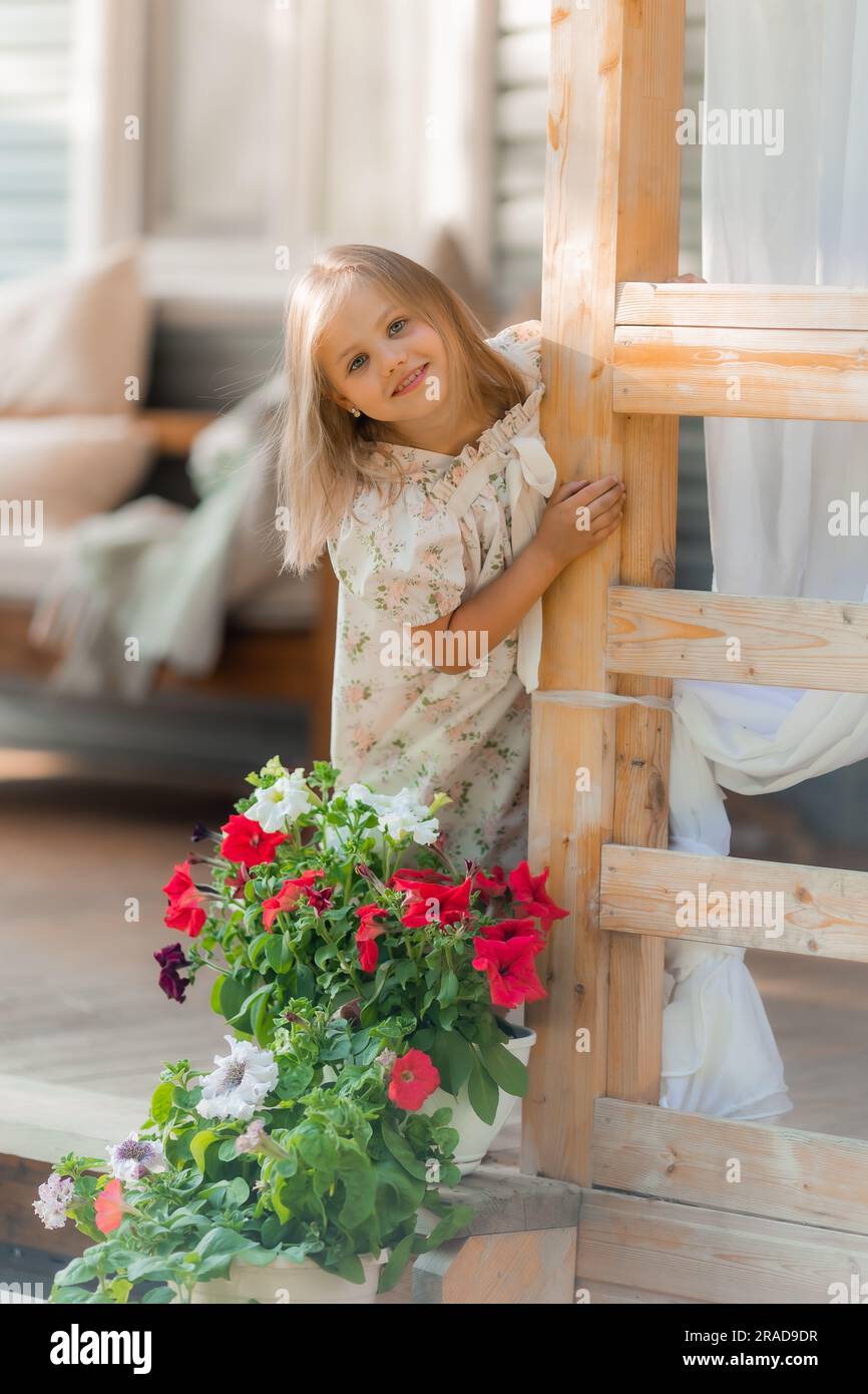 la bambina con i capelli biondi annaffia i fiori sulla veranda d'estate al tramonto. Foto di alta qualità Foto Stock