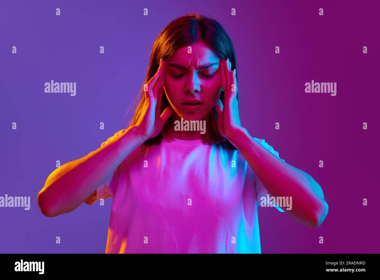 Ritratto di una giovane ragazza che tiene le mani sulla testa, ha mal di testa, viso premuroso su sfondo viola sfumato con luce al neon Foto Stock