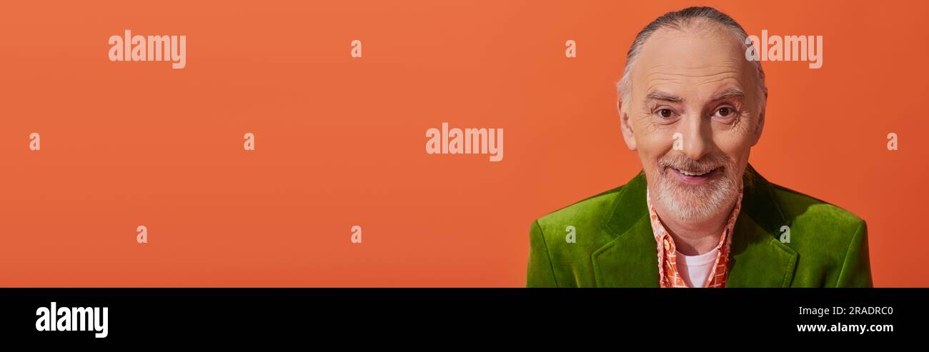 ritratto di un modello senior alla moda e allegro con capelli e barba grigi, con un blazer in velluto verde e un sorriso alla fotocamera su sfondo arancione vivace, Foto Stock