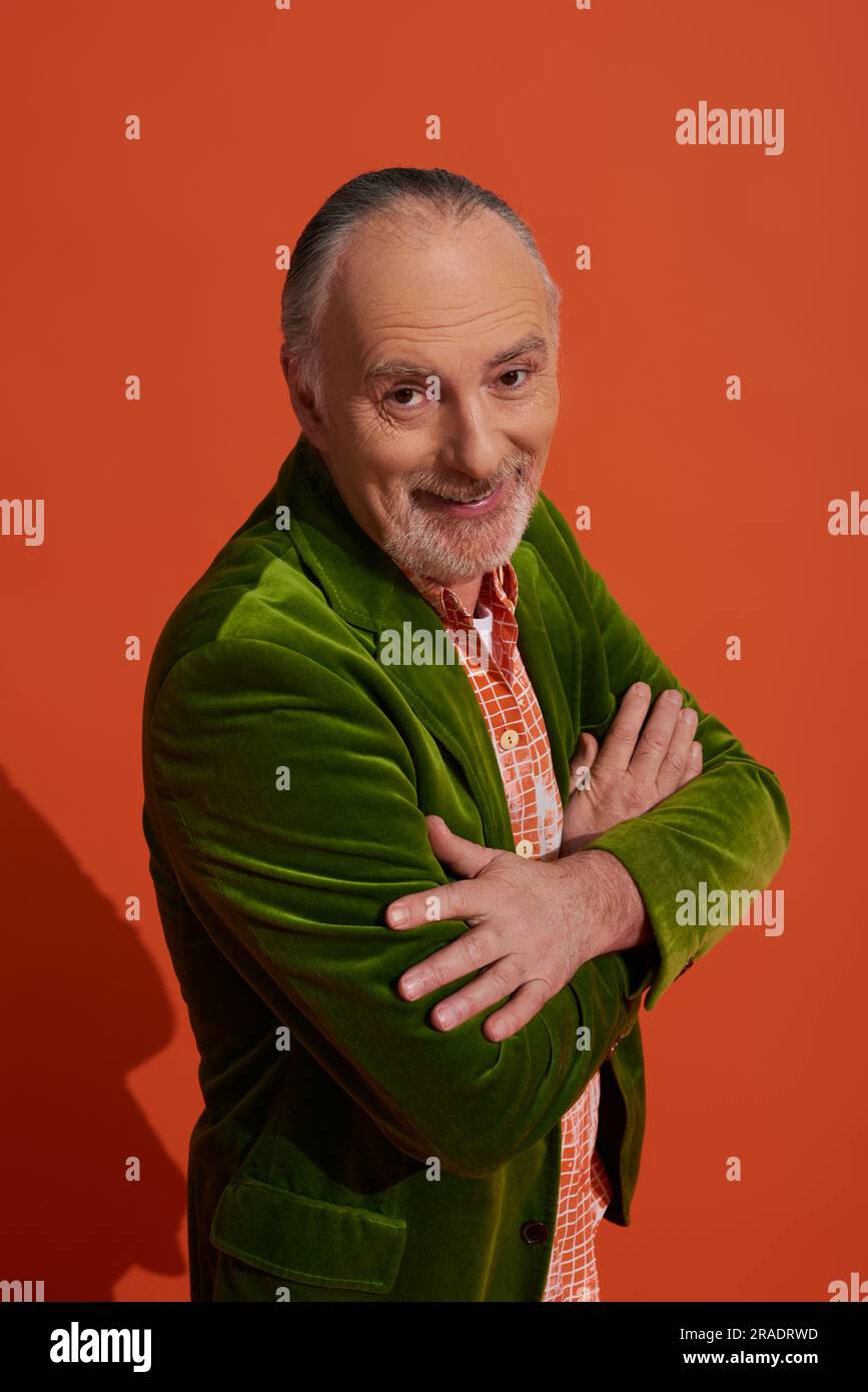 uomo anziano barbuto ottimista e carismatico in piedi con braccia piegate e sorridente davanti alla fotocamera su sfondo rosso arancione, look alla moda, stile personale, Foto Stock