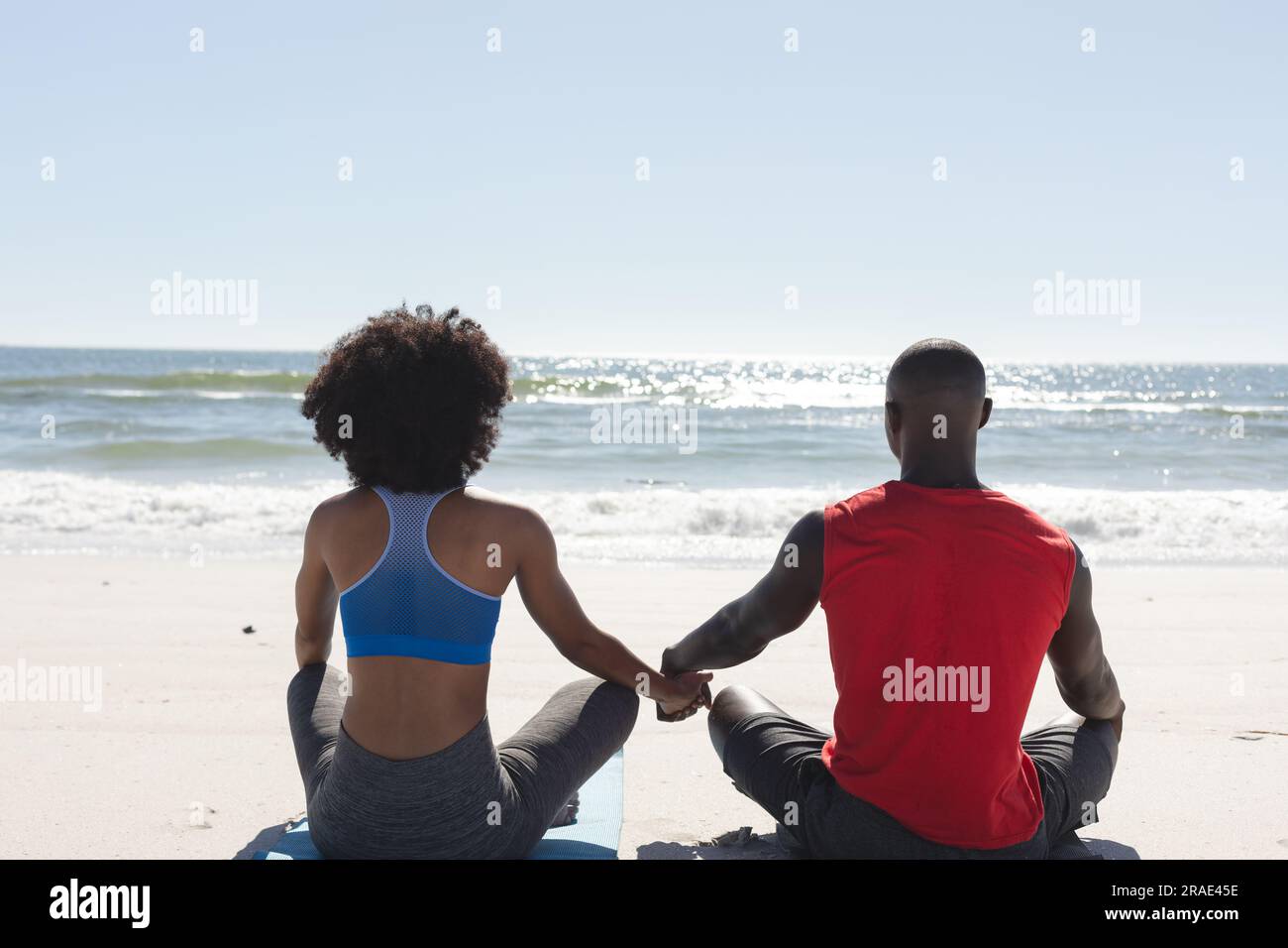Vista posteriore di una coppia afroamericana che pratica meditazione yoga e tiene le mani sulla spiaggia soleggiata Foto Stock