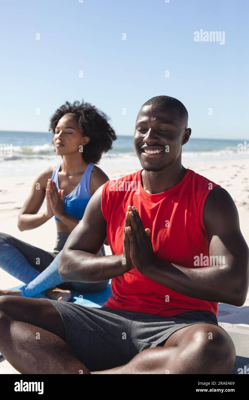 Coppia afro-americana felice e in forma che pratica meditazione yoga seduto sulla spiaggia soleggiata Foto Stock