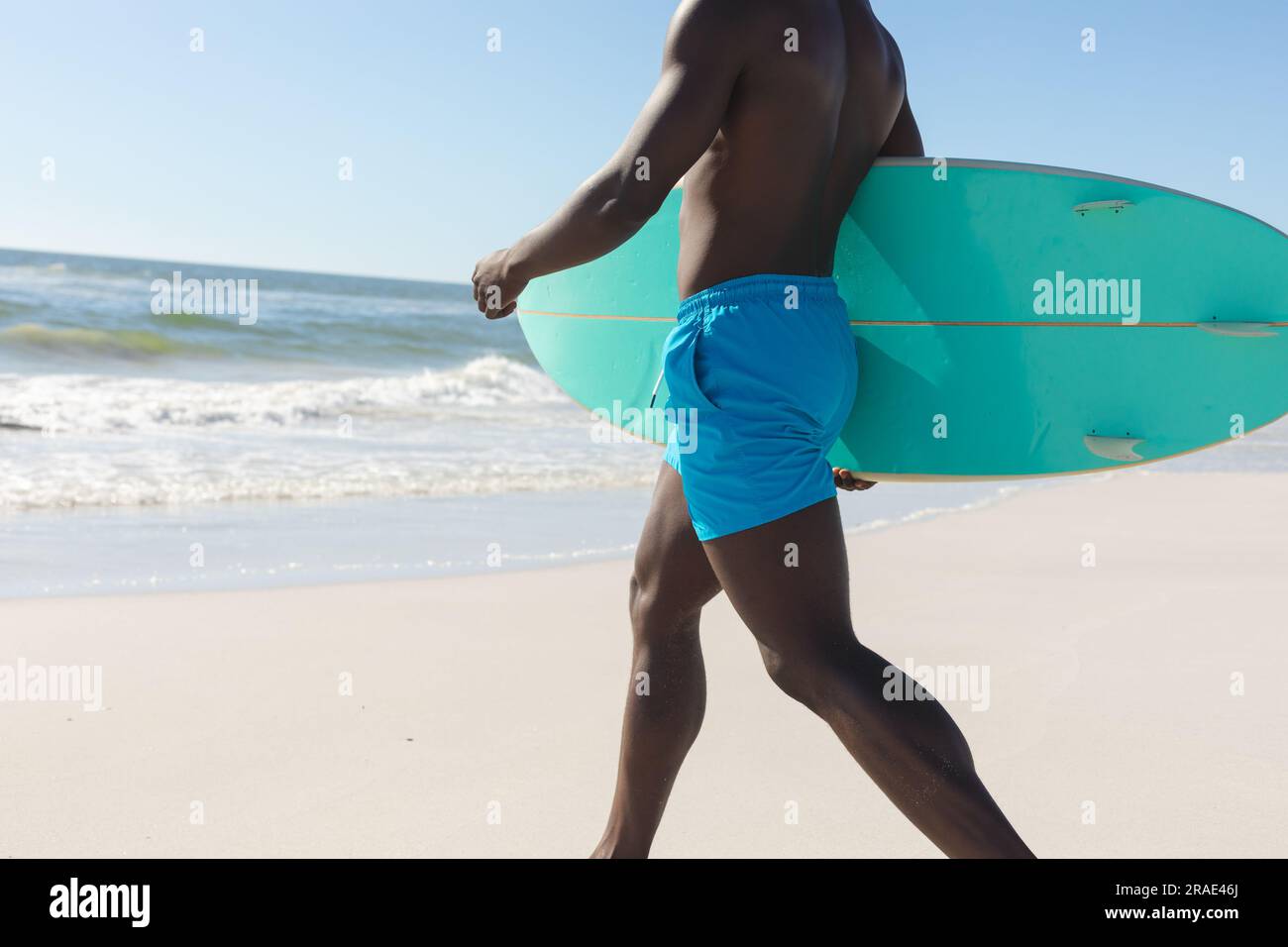 Sezione centrale di uomo afro-americano in forma che trasporta tavole da surf che cammina sulla spiaggia soleggiata fino al mare Foto Stock