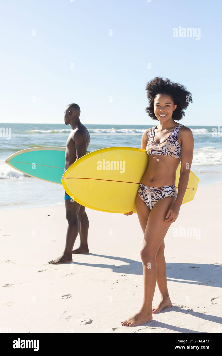 Ritratto di una donna afroamericana felice che tiene una tavola da surf in piedi sulla spiaggia soleggiata con un surfista maschio Foto Stock