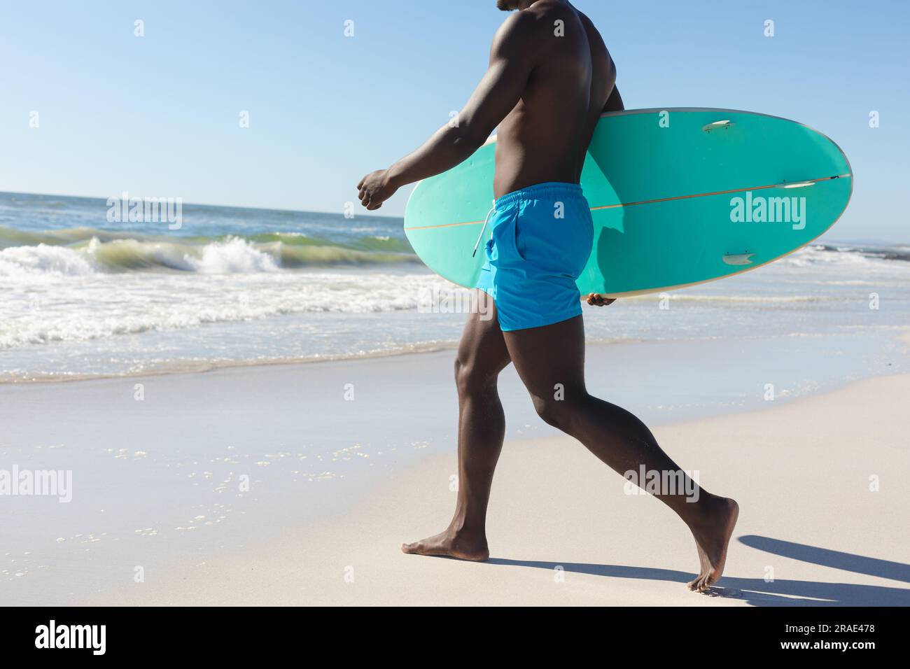 Sezione bassa di uomo afro-americano che trasporta tavole da surf che cammina sulla spiaggia soleggiata fino al mare, spazio fotocopie Foto Stock